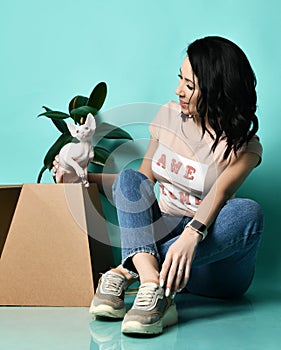 Female in beige t-shirt, blue jeans, sneakers. Smiling, sitting on floor near a paper box with Sphynx, posing on blue background