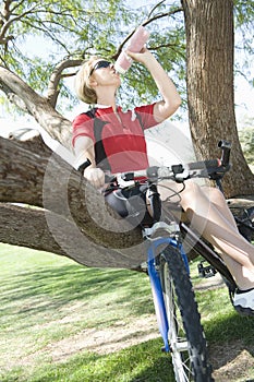 Female Bicyclist Sitting On Tree While Drinking Water