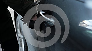 Female with black leather glove trying to break in car by using metallic stick.