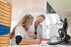 Female blonde pharmacist helps and assists her young aprrentice trainee during studies of medical books