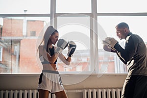 Female boxer practicing hits with her personal trainer in a boxing studio.