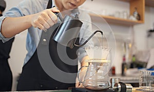 A female cafe operator wearing an apron pours hot water over roasted coffee grounds to prepare coffee for customers in the shop