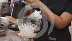 A female cafÃ© operator wearing an apron pours hot water over roasted coffee grounds to prepare coffee for customers in the shop