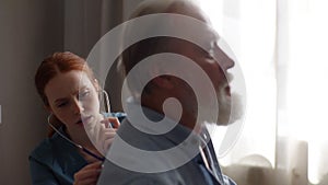 Female cardiologist doctor examining lungs of elderly senior male patient at home using stethoscope.