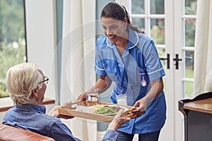 Female Care Worker In Uniform Bringing Meal On Tray To Senior Man Sitting In Lounge At Home
