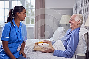 Female Care Worker In Uniform Bringing Senior Man At Home Breakfast In Bed On Tray