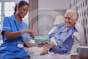 Female Care Worker In Uniform Helping Senior Man At Home In Bed With Medication