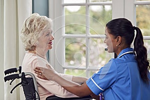 Female Care Worker In Uniform Talking With Senior Woman Sitting In Wheelchair In Care Home Lounge