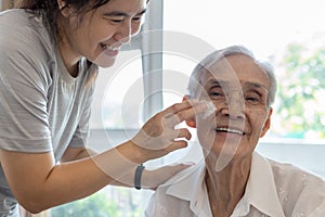 Female caregiver assisting to apply sunscreen lotion on the face of senior woman,granddaughter using skin care cream for elderly