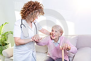 Female caregiver helping senior woman get up from couch in living room. Smiling nurse assisting senior woman to get up. Caring