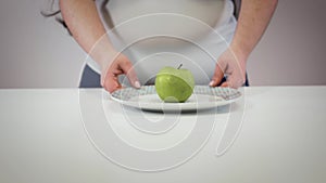 Female Caucasian hands taking plate with apple and showing folds on belly. Fat young woman having overweight problems