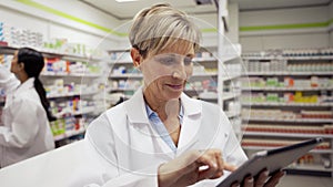 Female caucasian pharmacist typing on digital tablet emailing scripts to customers wearing white coat standing in