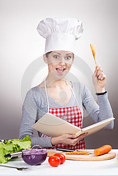 Female chef with the cookbook and a wooden spoon on gray background