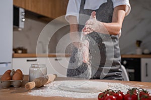 Female chef in a gray apron claps her hands with splashes of white flour on the background of the kitchen