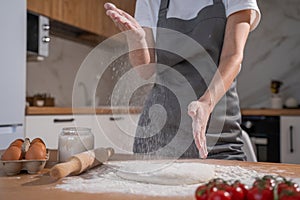 Female chef in a gray apron claps her hands with splashes of white flour on the background of the kitchen