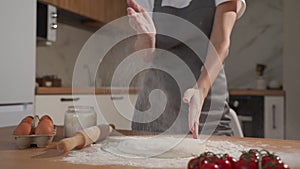 Female chef in a gray apron claps her hands with splashes of white flour on the background of the kitchen