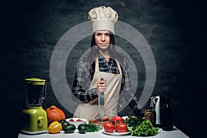 Female chef holds a knife and posing near a table with fresh vegetables.