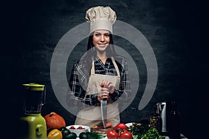 Female chef holds a knife and posing near a table with fresh vegetables.