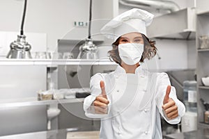 Female chef in mask showing thumbs up at kitchen