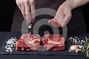 A female chef sprinkles sea salt with two fresh raw ribeye steaks from marbled beef on a dark background. Nearby is a mixture of p