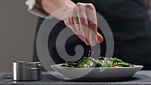 A female chef in a white uniform and a black apron in the restaurant kitchen. The cook sprinkles the salad with sesame