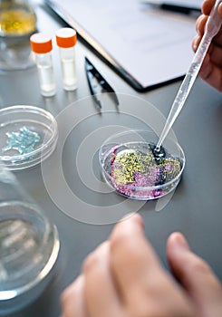 Female chemist adding drop of liquid from from pipette over petri dish with mix of glitters in lab