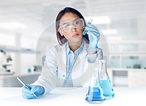Female chemist wearing lab coat, glasses and gloves holding a test tube and writing sample data in chemical laboratory