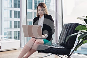 Female chief economist analyzing data using laptop sitting on armchair in modern office