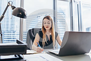 Female chief executive sitting at her desk taking notes in datebook writing with pen and using her computer in modern photo