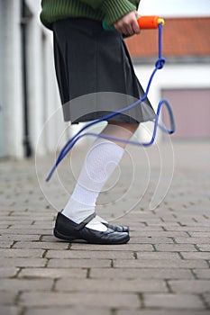 Female child with grey skirt and green coat, shot side on from the waist down while skipping with a blue skipping rope, on cobbles