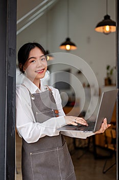 A female coffee shop owner smiling and leaning on the wall with a laptop in her hand