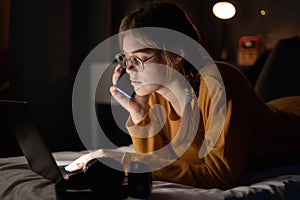 Female college student calling her friends, lying on bed working on laptop at night in dormitory.