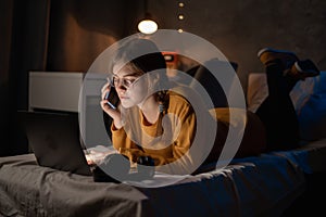 Female college student talking on mobile phone, lying on bed working on laptop at night in dormitory.