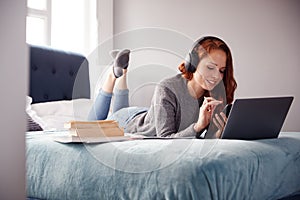 Female College Student Wearing Headphones Lies On Bed In Shared House With Mobile Phone Using Laptop