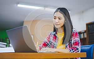 Female college student working on laptop and searching for books to study, make report, find useful information in college room.