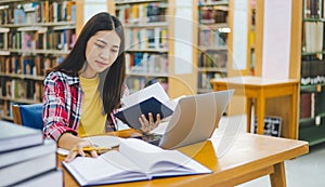 Female college student working on laptop and searching for books to study, make report, find useful information in college room.