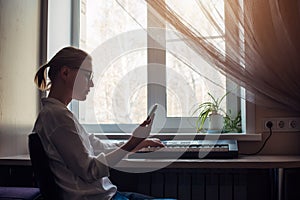 Female composer plays a synthesizer sitting at home near the window. Young girl in glasses and a white shirt makes music, reading