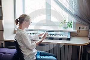 Female composer plays a synthesizer sitting at home near the window. Young girl in glasses and a white shirt makes music, reading