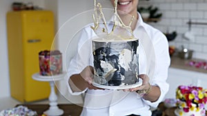 Female confectioner presenting decorated cake in her kitchen, holding it with outstretched hands. Aimed focus of cake