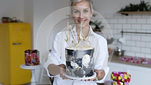 Female confectioner in white shirt presenting modern decorated black and white cake in her kitchen. Yellow fridge and