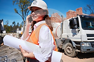 Female construction worker in hardhat holding blueprints and talking phone