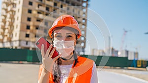 Female construction worker in overalls and medical mask recording audio message on smartphone on background of house