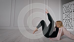 Female contemporary dancer is training in dance hall alone, sitting on floor, moving legs up
