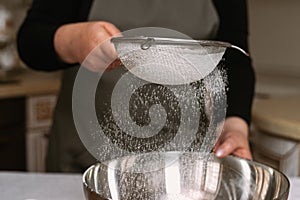 Female cook in apron sifts flour in a metal bowl. Preparing to create a dough for baking or sculpting convenience foods