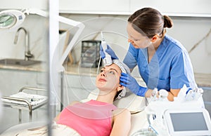 Female cosmetologist performing anti-aging face phototherapy procedure, shrink pores and repair damaged skin for young