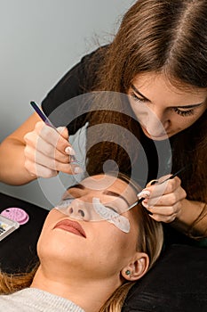 Female cosmetologist with tweezers attaches artificial eyelashes to female client at salon.