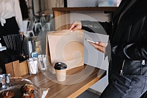 Female customer hold phone for contactless payment and take away package with food and cup of coffee during quarantine