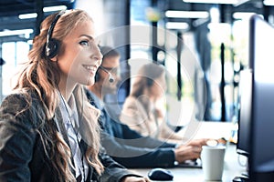 Female customer support operator with headset and smiling, with collegues at background.