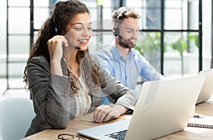 Female customer support operator with headset and smiling, with collegues at background.