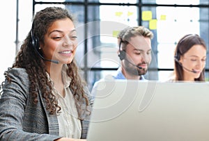 Female customer support operator with headset and smiling, with collegues at background.